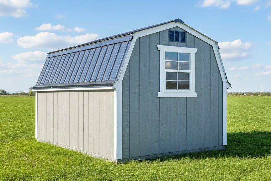 A small gray and white barn-style shed with a metal roof, shown outdoors on a gravel surface with buildings in the background.