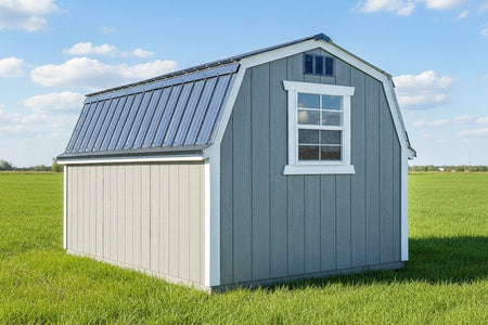A small gray and white barn-style shed with a metal roof, shown outdoors on a gravel surface with buildings in the background.